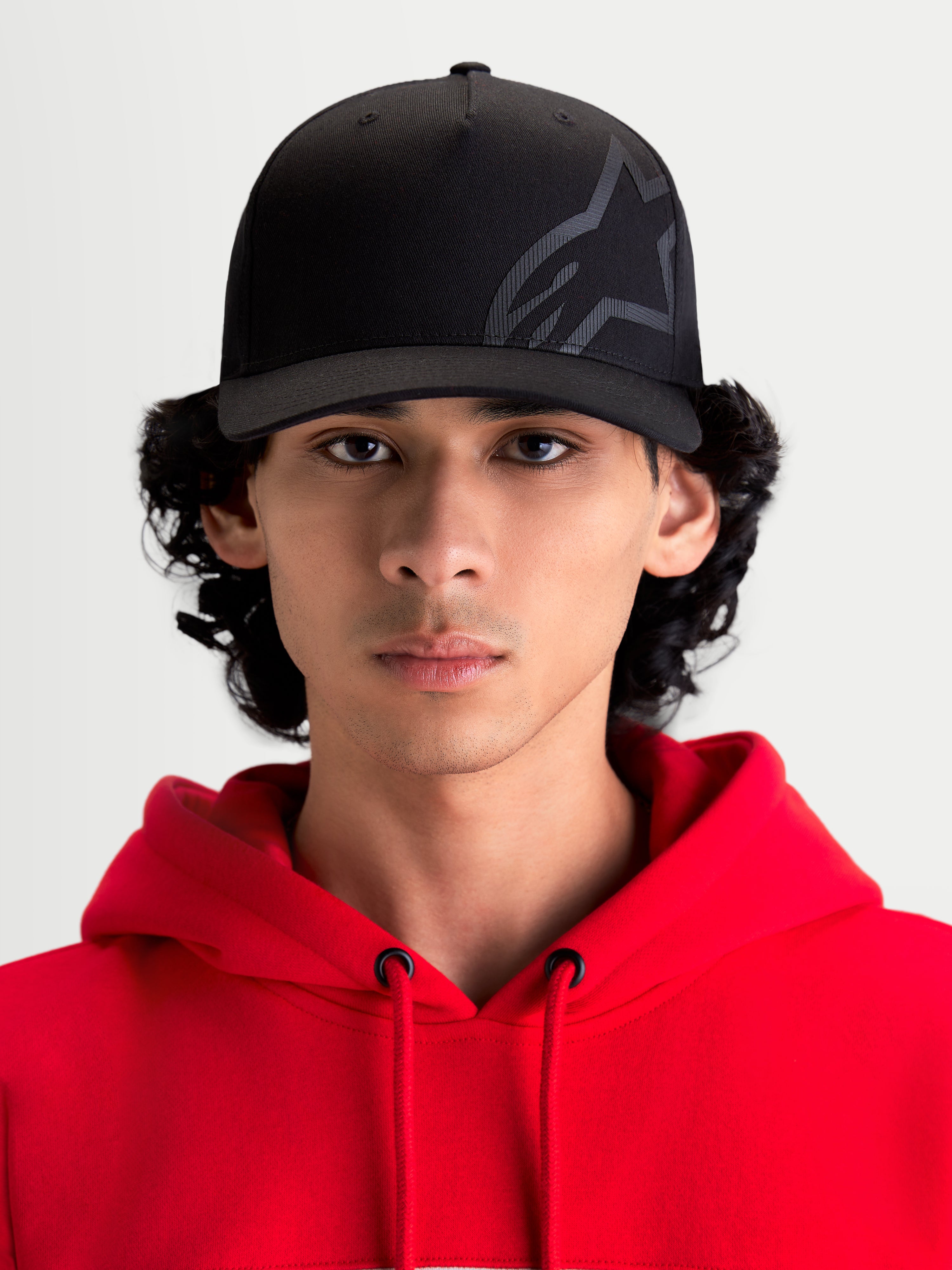 Alpinestars Maze keps, motorsportinspirerad sportkeps, svart, slitstark twill, strukturerad krona, böjd skärm och texturerad Astars Corp Shift-logotyp, frontvy på modell i röd huvtröja.