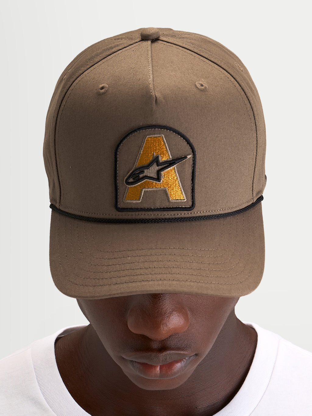 Alpinestars Acme Snapback keps, brun vardagskeps med böjd skärm, broderad Astars-logotyp framtill och svart repkant, bars av en modell