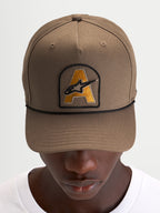 Alpinestars Acme Snapback keps, brun vardagskeps med böjd skärm, broderad Astars-logotyp framtill och svart repkant, bars av en modell