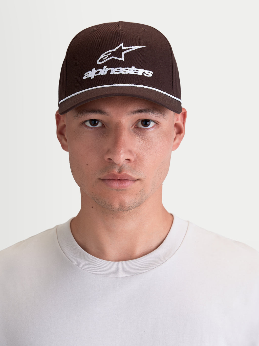 Alpinestars Combo Logo snapback-keps, sportswear-keps med böjd skärm, brun och vit, slitstark bomullstwill med broderad logotyp, avslappnad motorsportstil, vy framifrån på modell