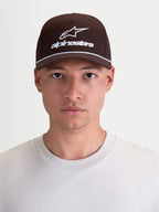 Alpinestars Combo Logo snapback-keps, sportswear-keps med böjd skärm, brun och vit, slitstark bomullstwill med broderad logotyp, avslappnad motorsportstil, vy framifrån på modell