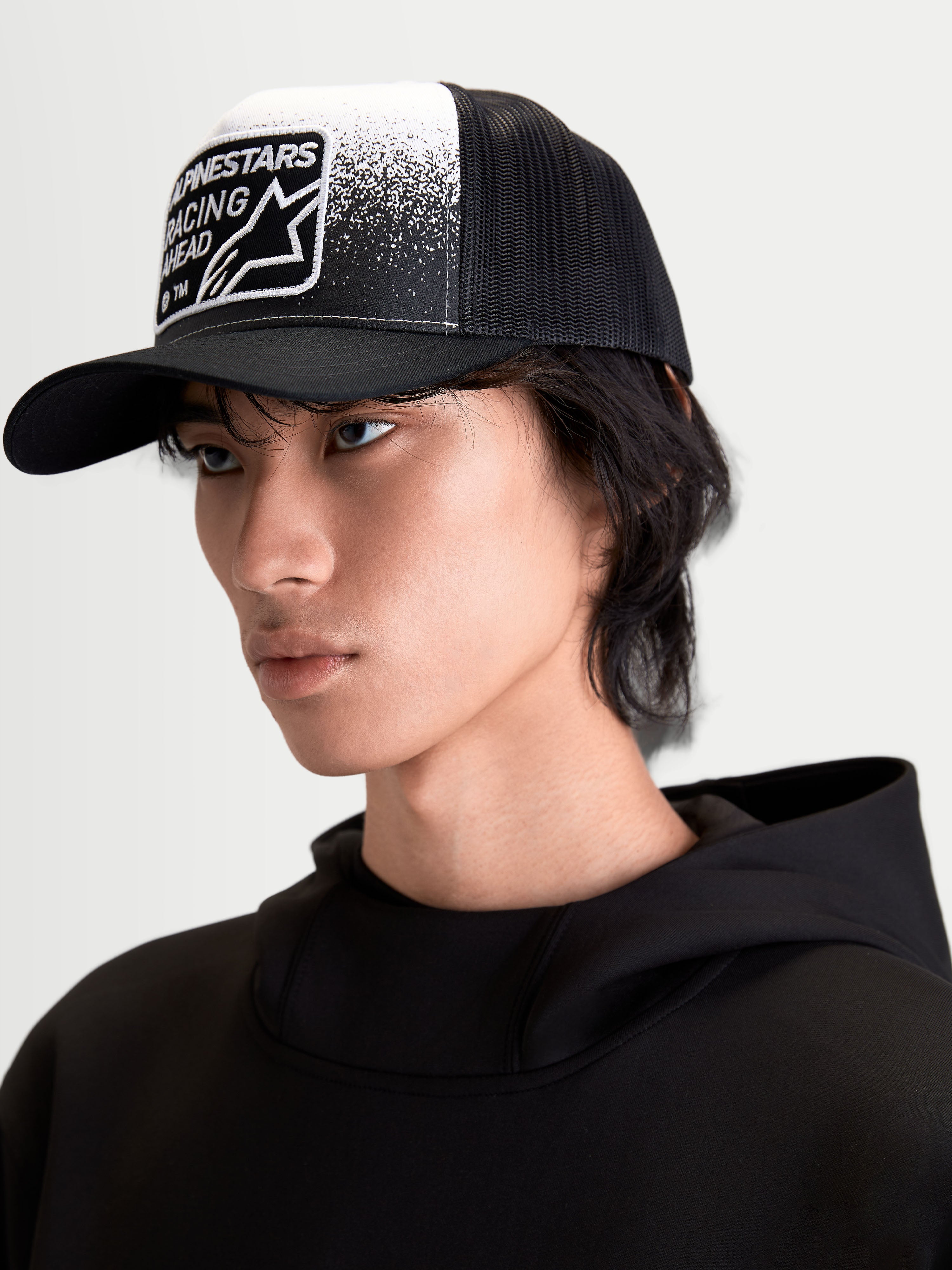 Alpinestars Gravel keps, trucker-keps för vardagsbruk på modell, svart och vit, med strukturerad krona, böjd skärm, ventilerande mesh-baksida och vävd patch med grustexturerad grafik och Alpinestars Racing Ahead-branding.