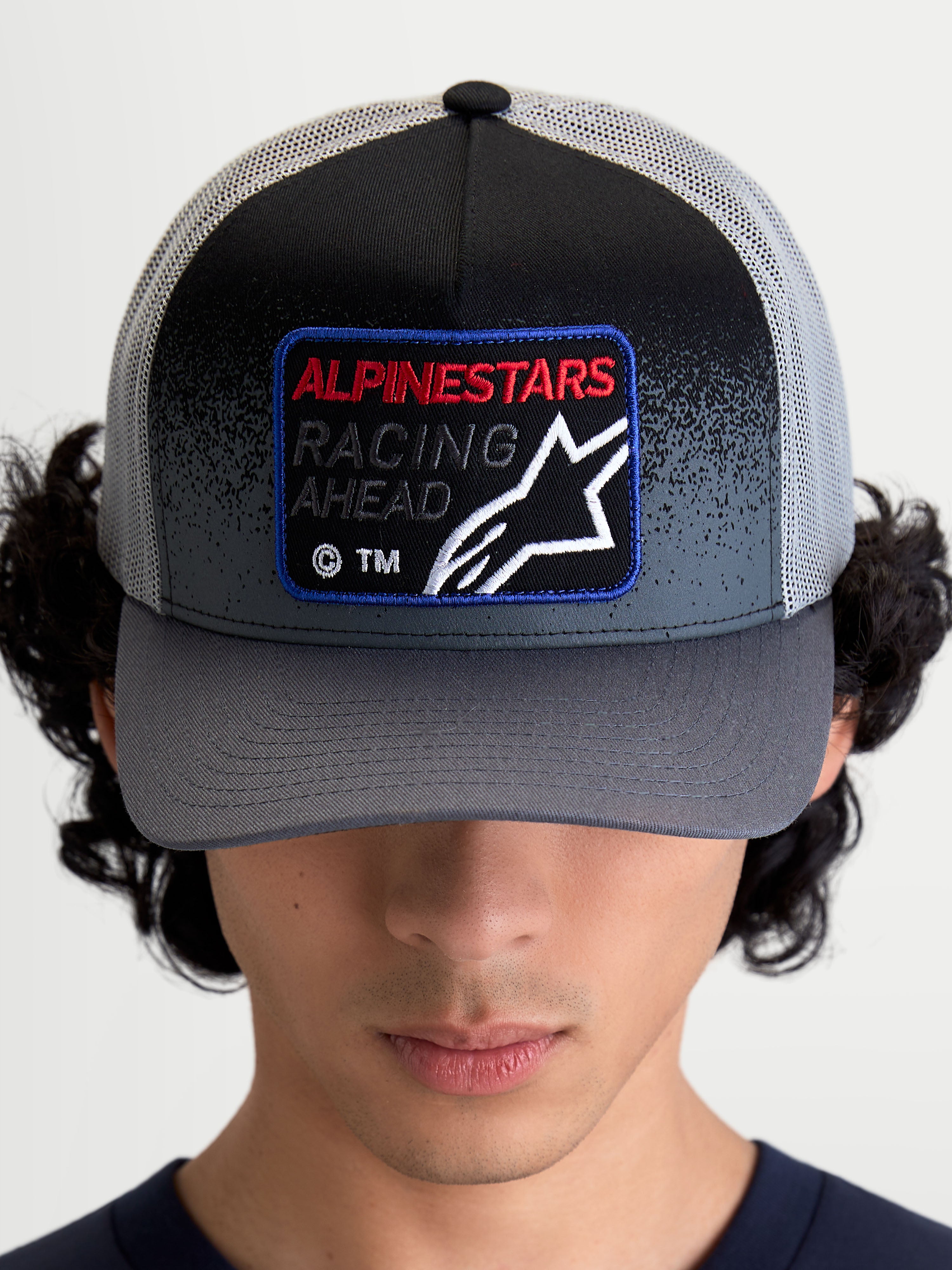 Alpinestars Gravel keps, mörkgrå och svart keps i truckermodell med böjd skärm och vävd frontpatch med texten Racing Ahead och Astars-logotyp, frontvy på modell.