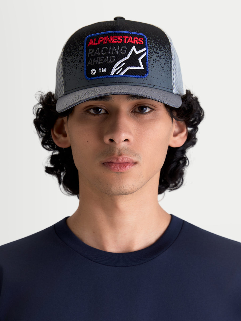 Alpinestars Gravel keps, truckerkeps med mesh-baksida för dam och herr, mörkgrå och svart, på modell. Har strukturerad krona, böjd skärm och vävd frontpatch med Alpinestars Racing Ahead-märkning och grustextur.