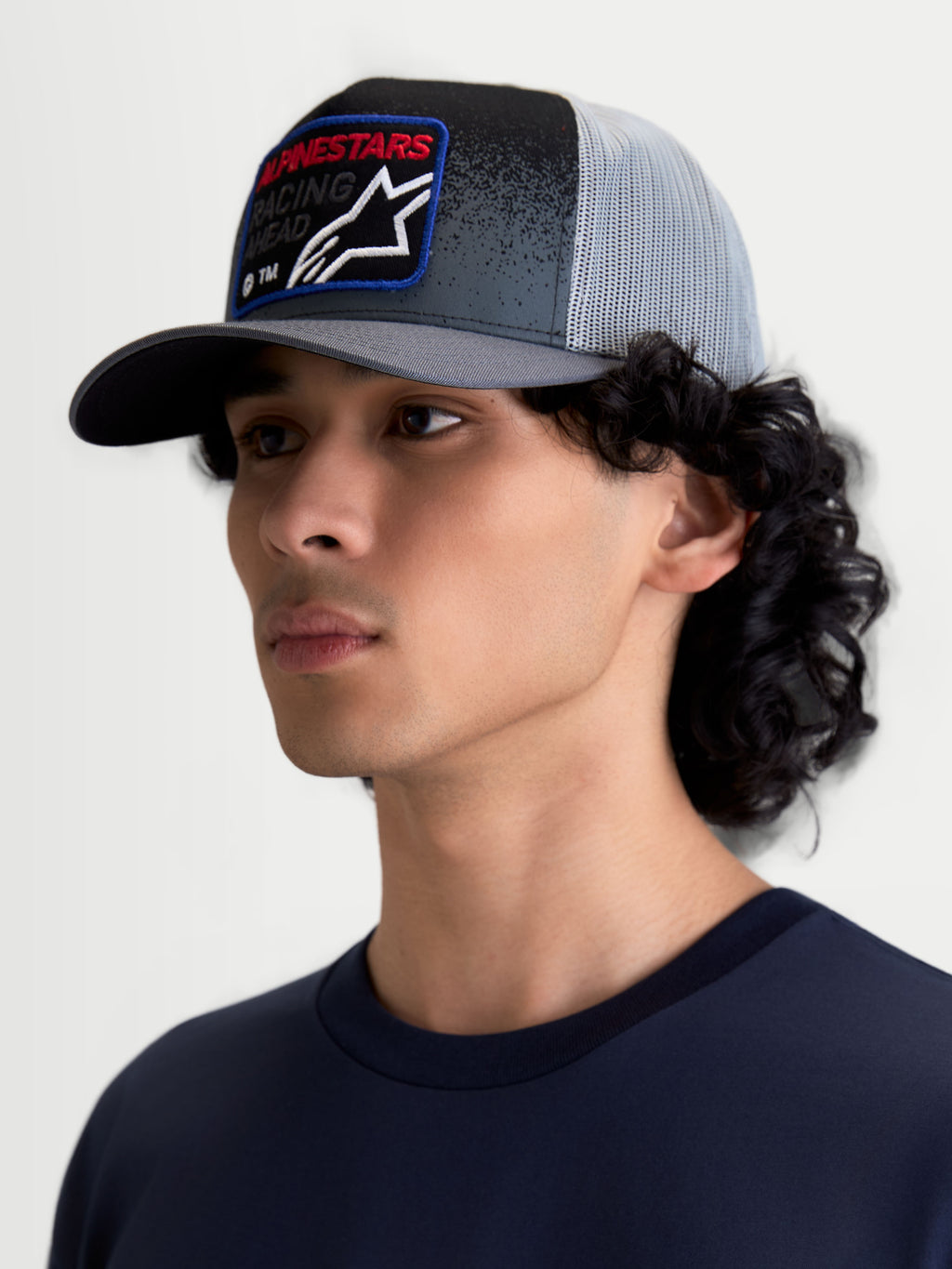 Alpinestars Gravel keps, truckerkeps i mörkgrått och svart med ljusgrå nätbaksida, böjd skärm och strukturerad krona, vävt märke på framsidan med röd text, motorsportsinspirerad sportswear på modell