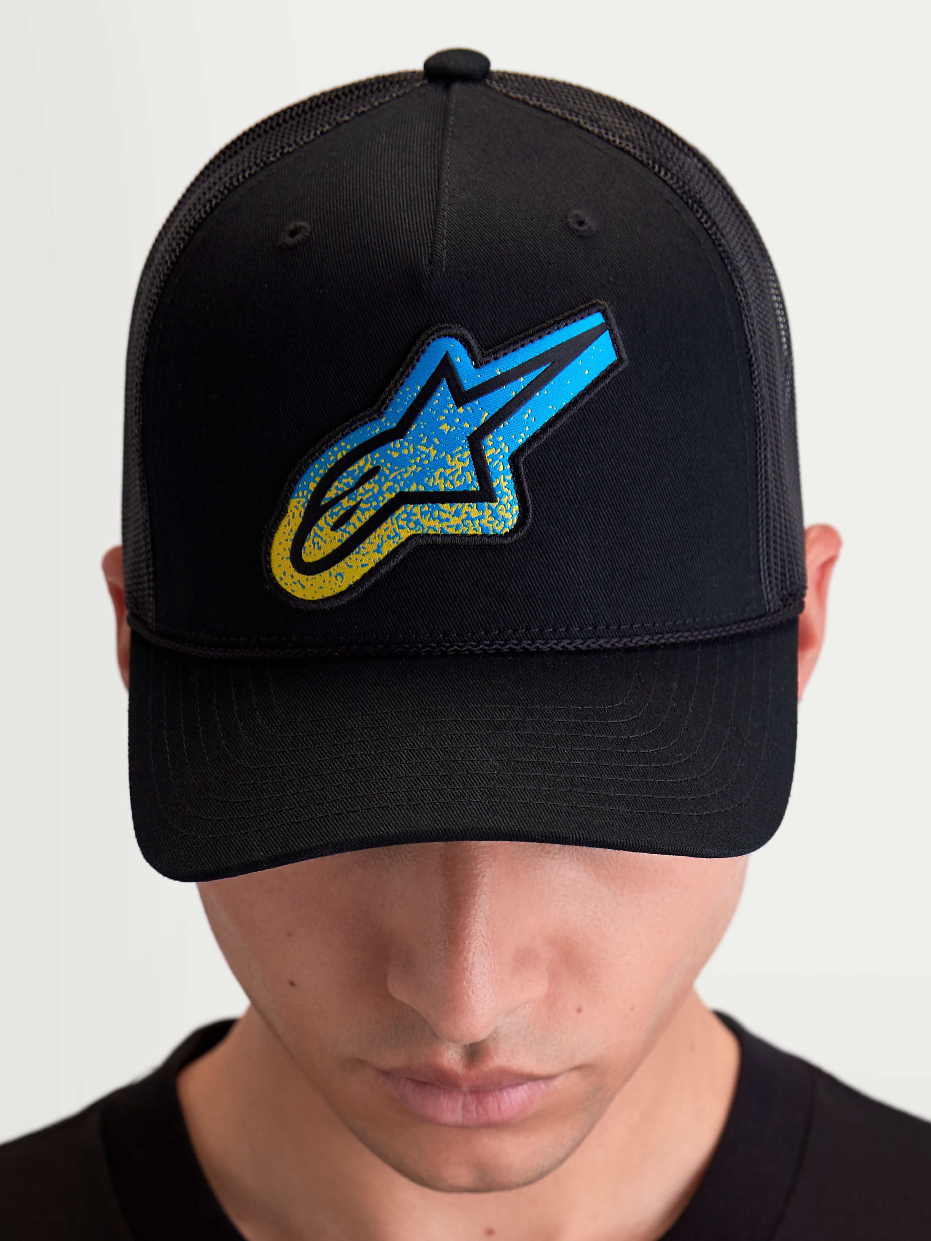 Alpinestars Meteorite keps, svart trucker-keps med blå och gul texturerad Astars-logotyp, strukturerad krona, böjd skärm och ventilerande mesh-baksida, bärs av en modell.