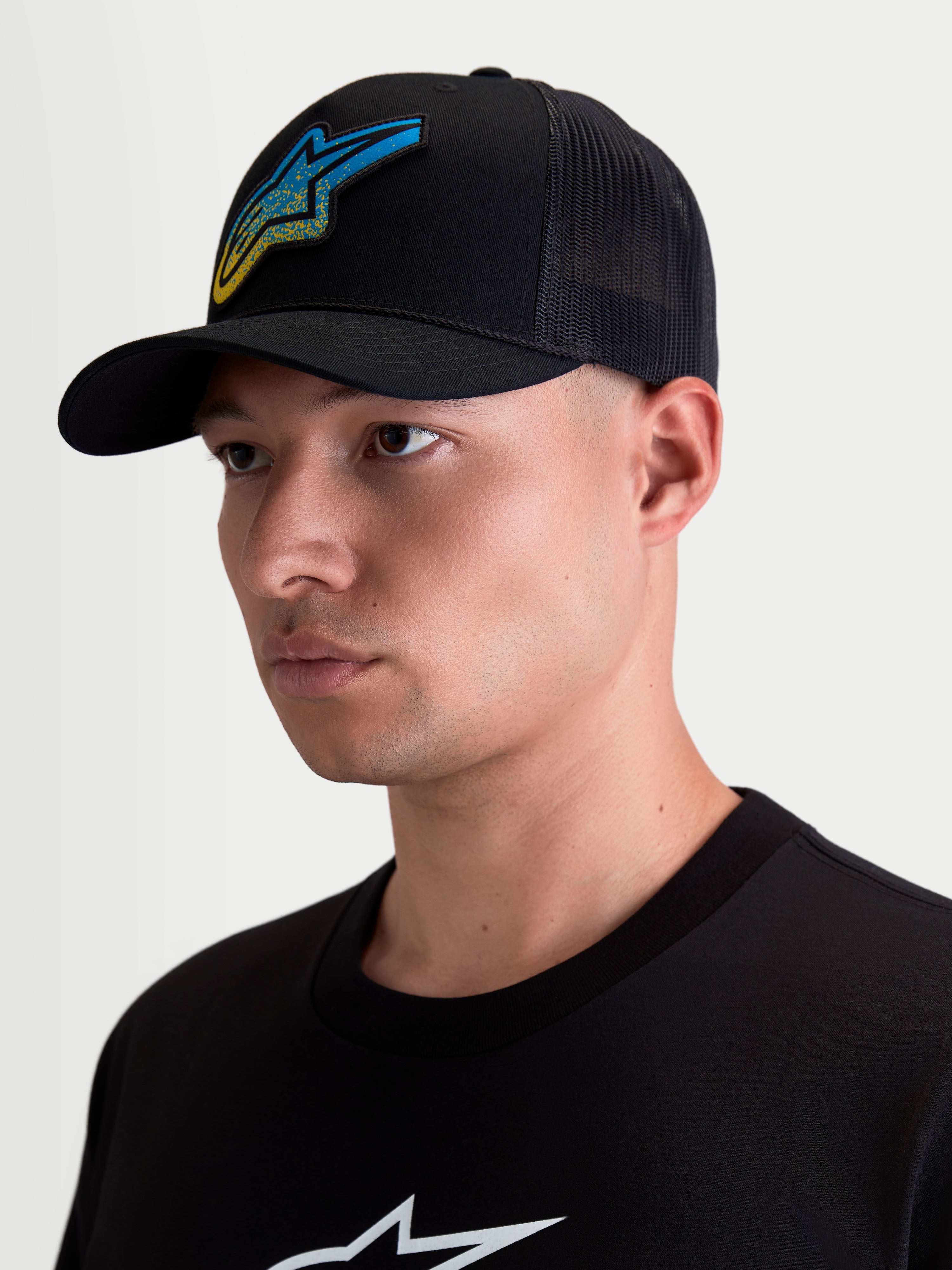 Alpinestars Meteorite keps, svart trucker-keps för herrar med blå och gul vävd Astars-logotyp i grustextur, böjd skärm, mesh-baksida och strukturerad krona, visas på modell.