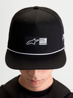 Alpinestars Label snapback-keps, svart och vit livsstilskeps i bomullstwill, strukturerad krona med platt skärm, vit snördetalj och vävd logotyplapp fram med Alpinestars-märkning, visad på modell.