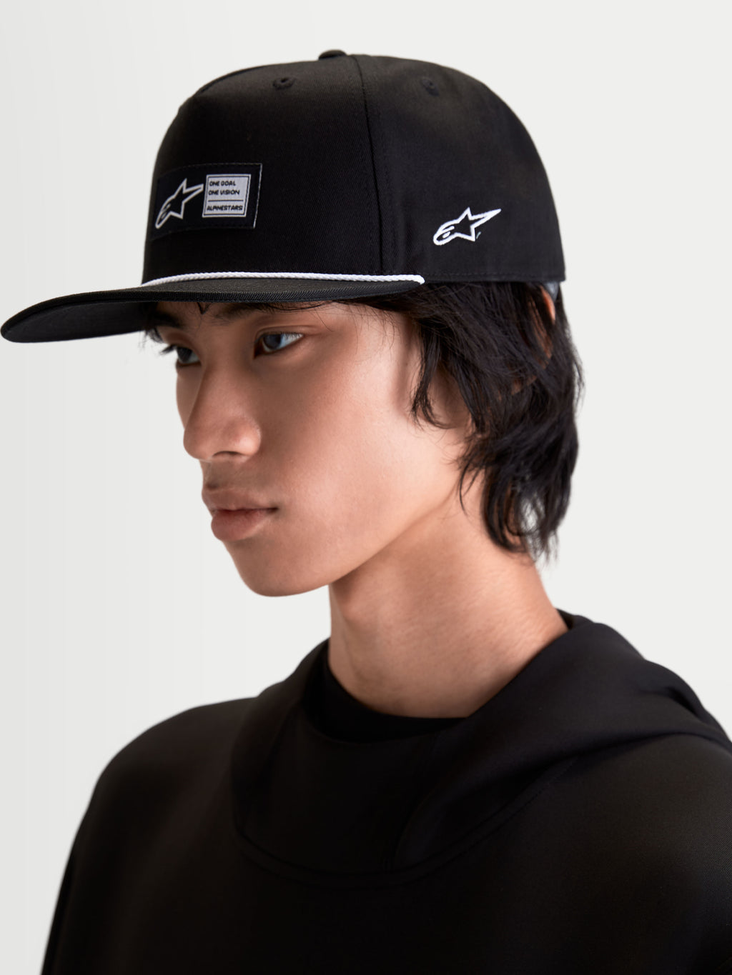 Alpinestars Label Snapback keps, svart sportkeps med platt skärm och strukturerad krona, vävd etikett framtill, vit dekorationssnörning och broderad logotyp på sidan, visas på modell för en modern motorsports-inspirerad look.