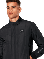 Alpinestars Precedent Jacka, quiltad sportjacka för herr i svart, närbild som visar den låga ståkragen, logomärke på bröstet och broderi på ärmen, med ett ikoniskt vinklat quiltmönster för en motorsport-inspirerad look.
