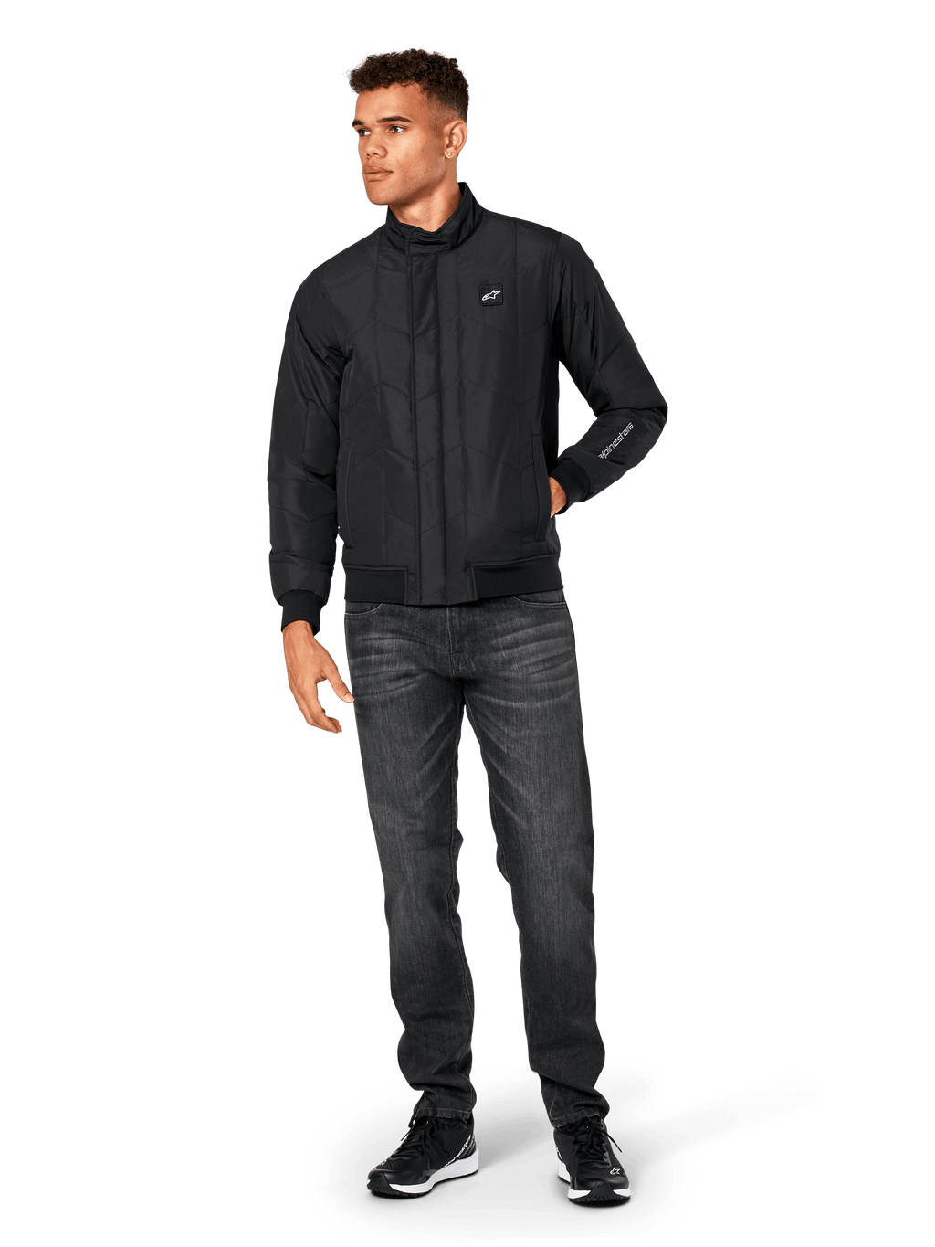 Alpinestars Precedent Jacket, quiltad sportjacka, svart, bärs med mörkgrå jeans och svarta sneakers, med ett vinklat quiltmönster, låg ståkrage, ribbstickade muddar och nederkant, vitt logomärke på bröstet och broderi på ärmen.