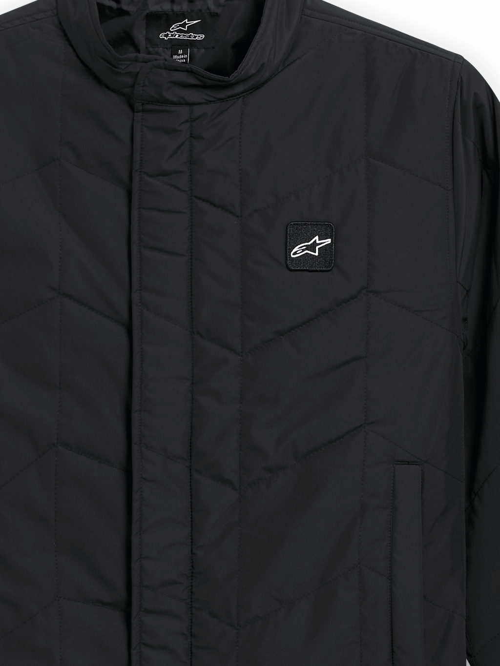 Alpinestars Precedent Jacka, quiltad sportswearjacka, svart, närbild som visar ett ikoniskt vinklat quiltmönster, låg ståkrage, täckt dragkedja framtill och ett litet logomärke på bröstet