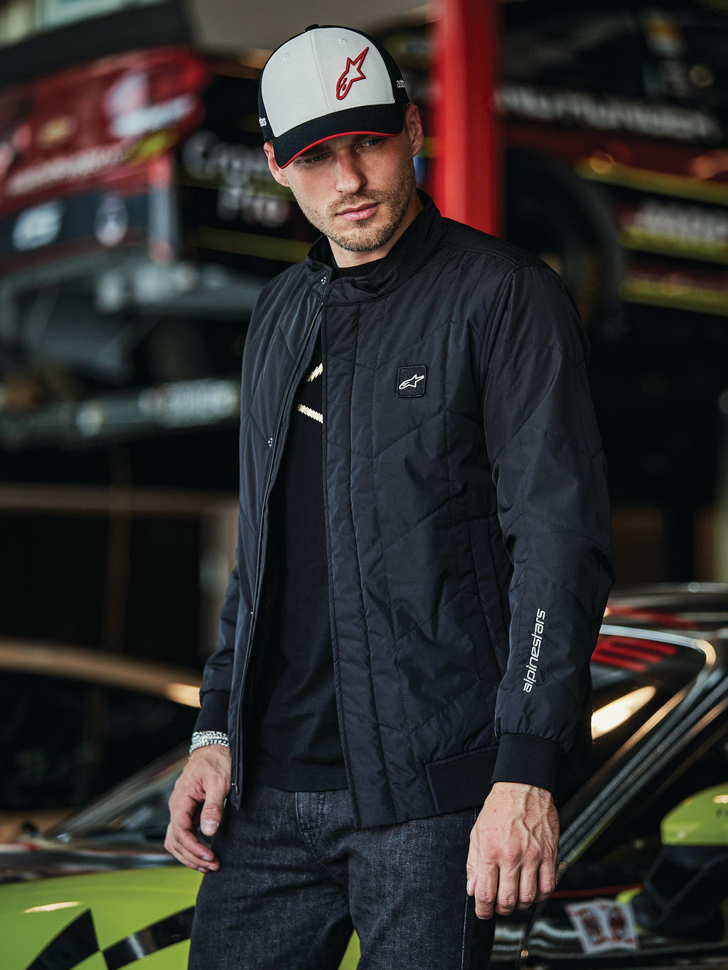 Alpinestars Precedent Jacket, svart quiltad jacka med Formula 1-stil, med ett ikoniskt vinklat quiltmönster, logomärke på bröstet och vitt broderi på ärmen, bärs tillsammans med en matchande Alpinestars-keps i en motorsportgarage-miljö.