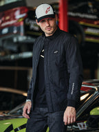 Alpinestars Precedent Jacket, svart quiltad jacka med Formula 1-stil, med ett ikoniskt vinklat quiltmönster, logomärke på bröstet och vitt broderi på ärmen, bärs tillsammans med en matchande Alpinestars-keps i en motorsportgarage-miljö.
