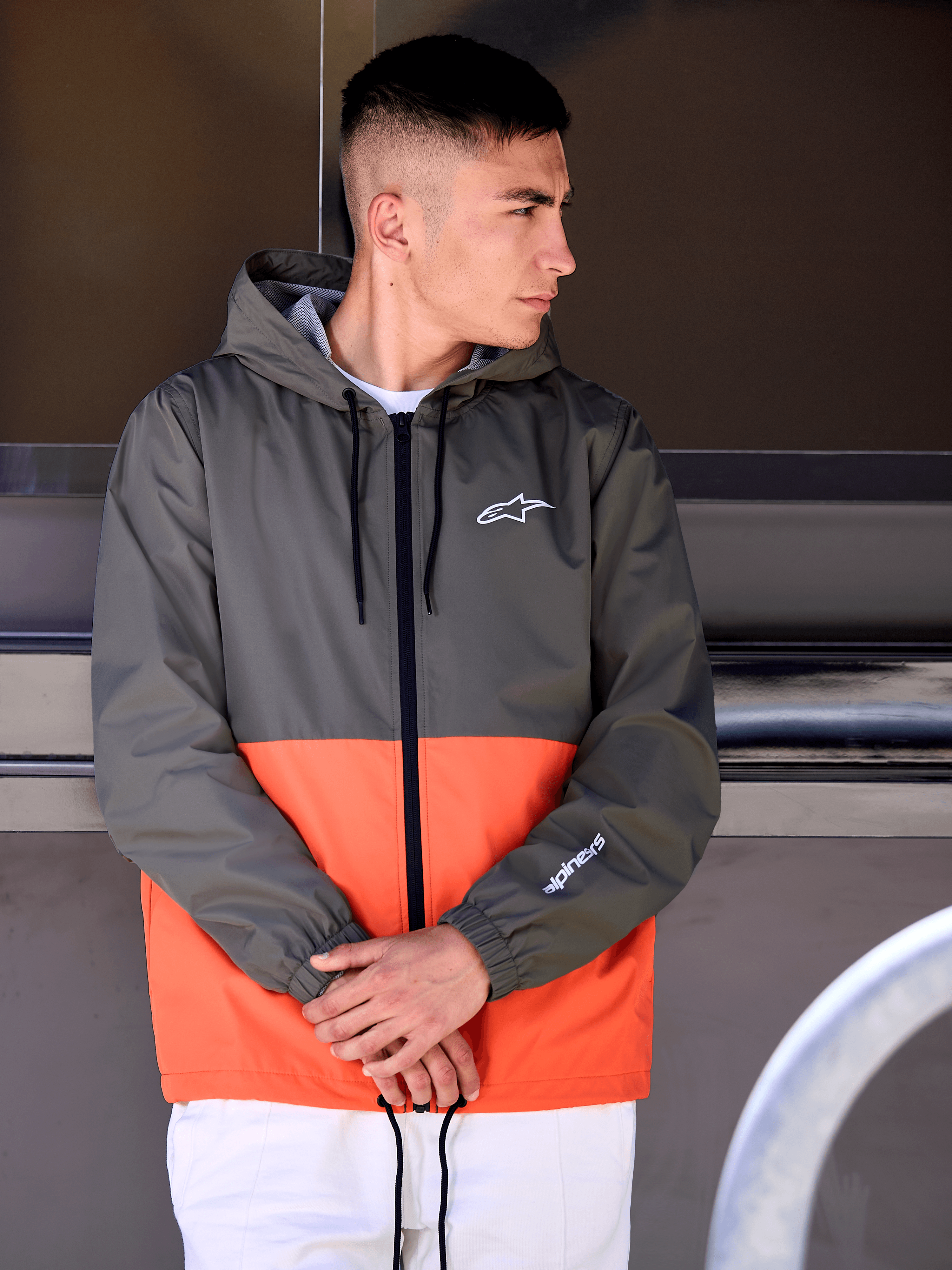 Man som bär Alpinestars Speeder vindjacka, blockfärgad design i militärorange och mörkgrönt, ytterplagg med huva och vita logotyper på bröst och ärm, kombinerat med vita byxor för en motorsportinspirerad livsstilslook