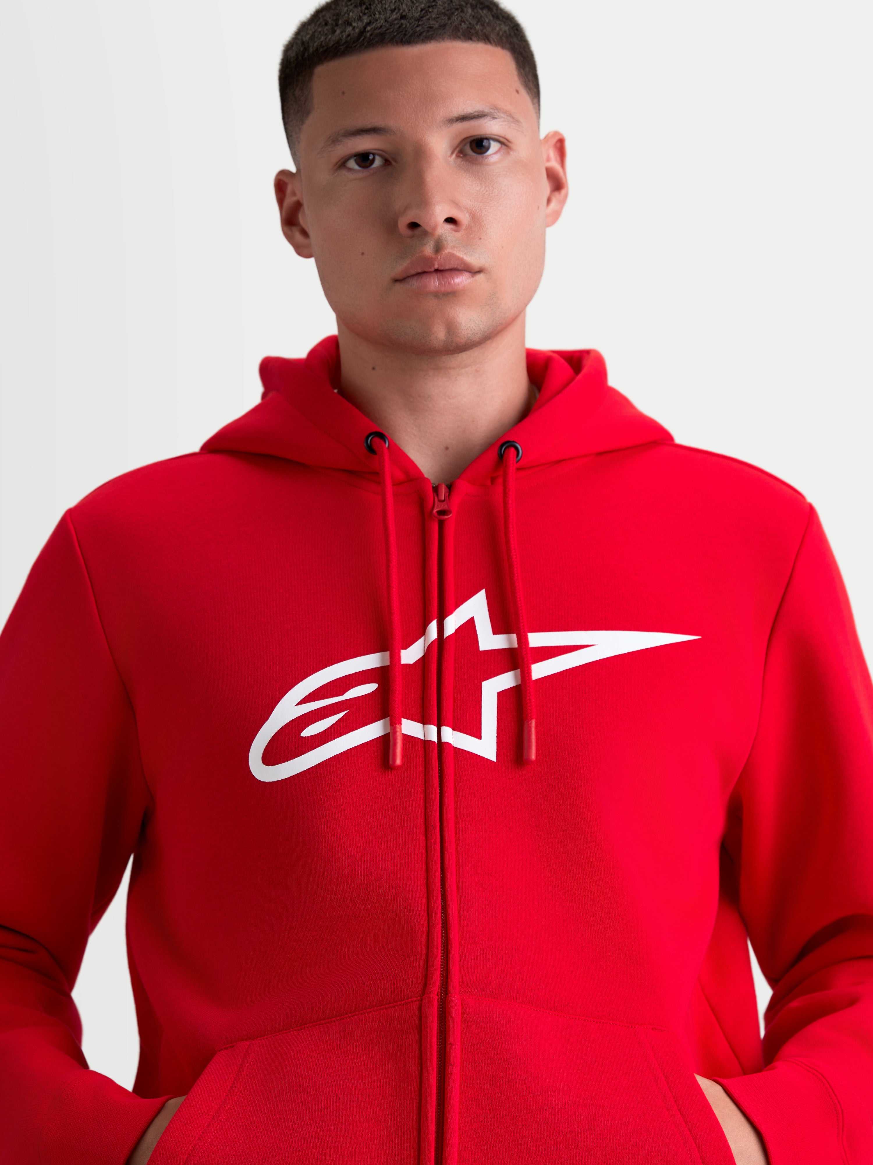 Alpinestars Ageless Zip V3 huvtröja, röd och vit sportswear-fleecehuvtröja med dragkedja, stor vit A-star-logotyp på bröstet, huva med justerbara dragskor och dold dragkedja framtill, motorsportinspirerad design på modell.