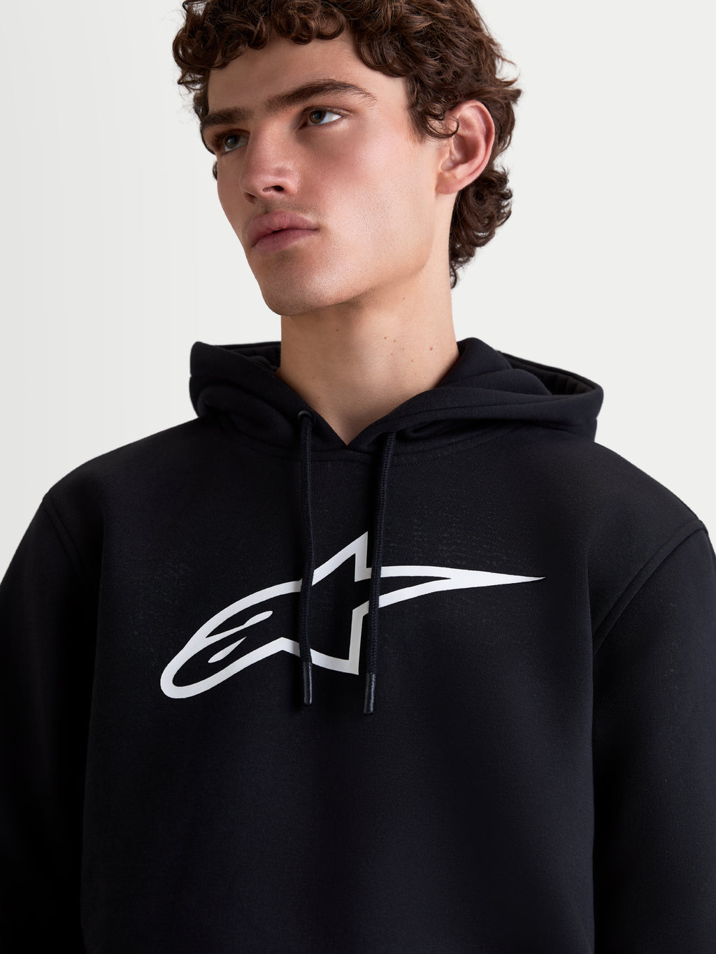 Alpinestars Ageless V3 huvtröja, sportig pullover i fleece för herrar, svart och vit, närbild på modell med stor vit logotyp på bröstet och huva med dragsko, mjuk fleecekvalitet för vardagskomfort.