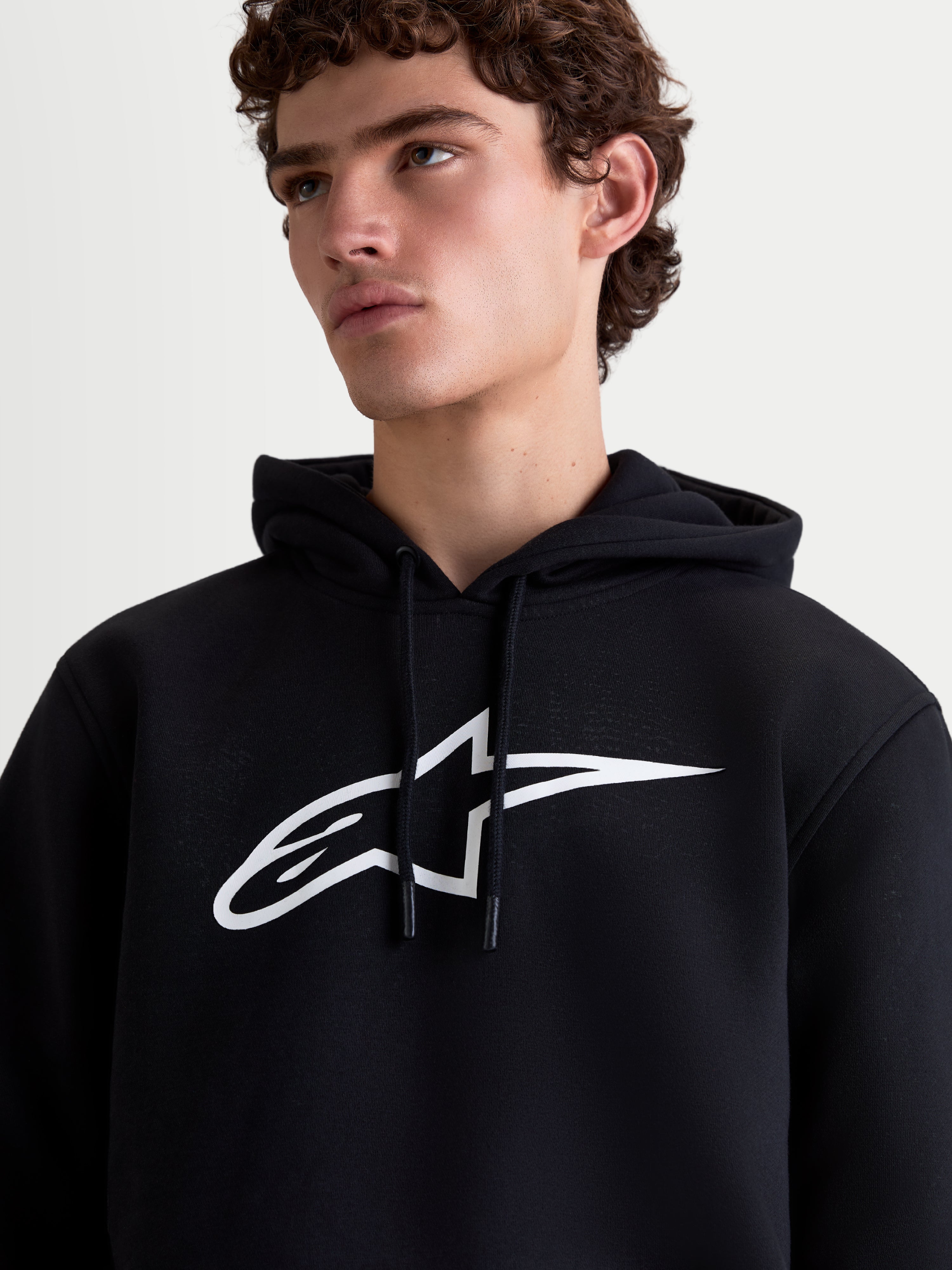 Alpinestars Ageless V3 huvtröja, sportig pullover i fleece för herrar, svart och vit, närbild på modell med stor vit logotyp på bröstet och huva med dragsko, mjuk fleecekvalitet för vardagskomfort.