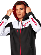 Alpinestars Sessions V3 Hoodie, svart, vit och röd, huvtröja med dragkedja bärs av en man med huvan uppe, röda Alpinestars-logotyper på bröstet och ärmarna, avslappnad motorsport-streetwear