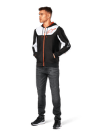 Alpinestars Sessions V3 Hoodie, vardaglig huvtröja med dragkedja fram, svart, vit och röd, bärs av en modell med mörka jeans och svarta sneakers, vita färgblockspaneler på bröstet och ärmarna, röda detaljer på dragkedja och dragsnören, stor bröstlogotyp, motorsportinspirerat streetwear i polyfleece