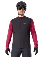 Alpinestars A-Dura Thermal Vest, svart isolerad cykelväst, bärs av en modell med en vinröd långärmad tröja, svart MTB-hjälm och solglasögon, med en vit logotyp på bröstet, lättviktigt termiskt skydd för mountainbike.