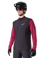 Alpinestars A-Dura Thermal Vest, svart, termisk cykelväst, bärs över en vinröd långärmad tröja, med en vit logotyp på bröstet, kombinerat med en svart MTB-hjälm och solglasögon, isolerande funktionslager för mountainbike