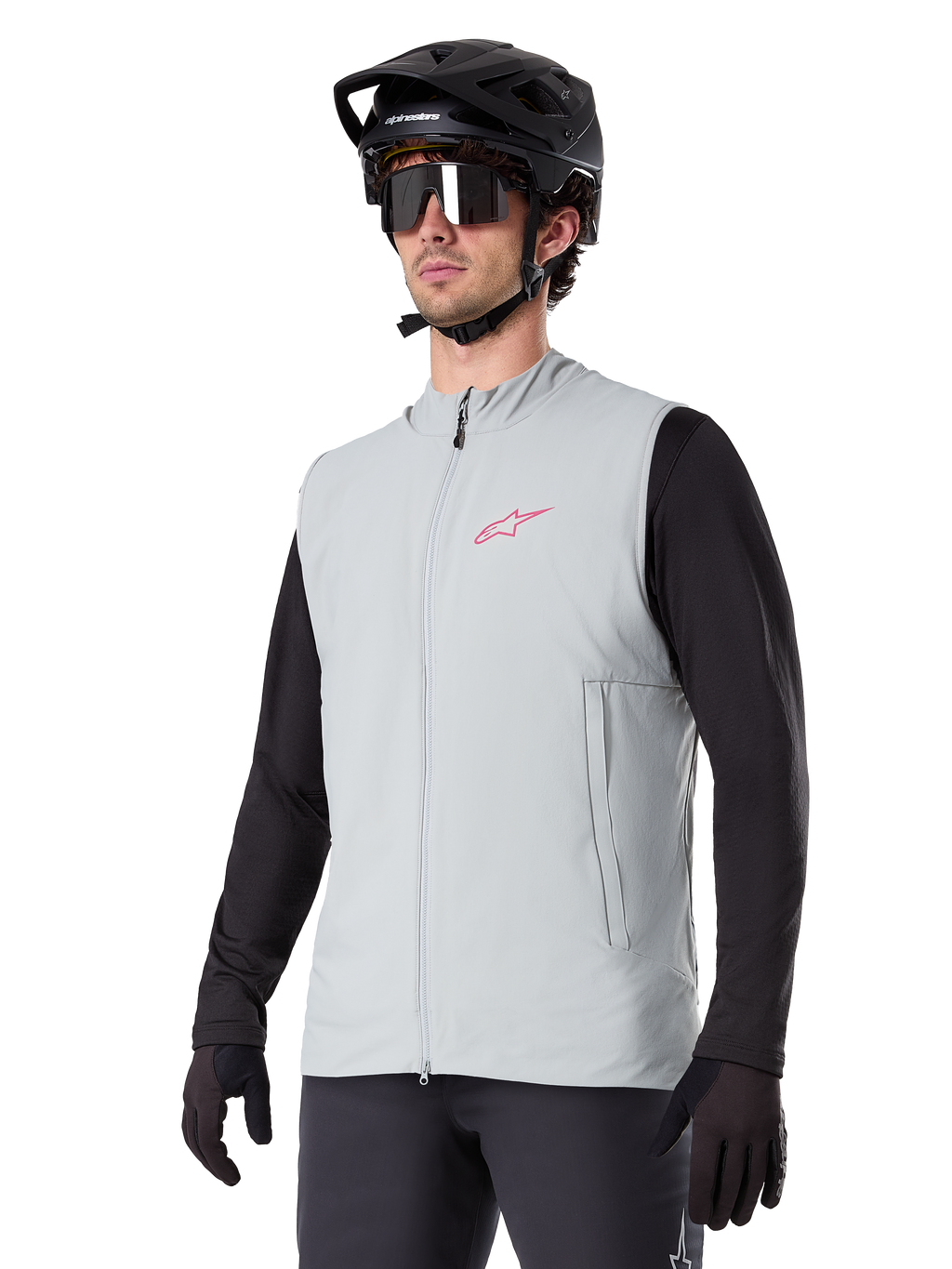 Alpinestars A-Dura Thermal Vest, titangrå, isolerad cykelväst, bärs över en svart långärmad tröja med en svart mountainbikehjälm och handskar, med en rosa logotyp på bröstet och vertikala sidofickor med dragkedja