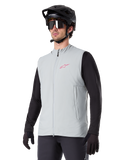 Alpinestars A-Dura Thermal Vest, titangrå, isolerad cykelväst, bärs över en svart långärmad tröja med en svart mountainbikehjälm och handskar, med en rosa logotyp på bröstet och vertikala sidofickor med dragkedja