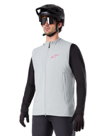 Alpinestars A-Dura Thermal Vest, titangrå, isolerad cykelväst, bärs över en svart långärmad tröja med en svart mountainbikehjälm och handskar, med en rosa logotyp på bröstet och vertikala sidofickor med dragkedja