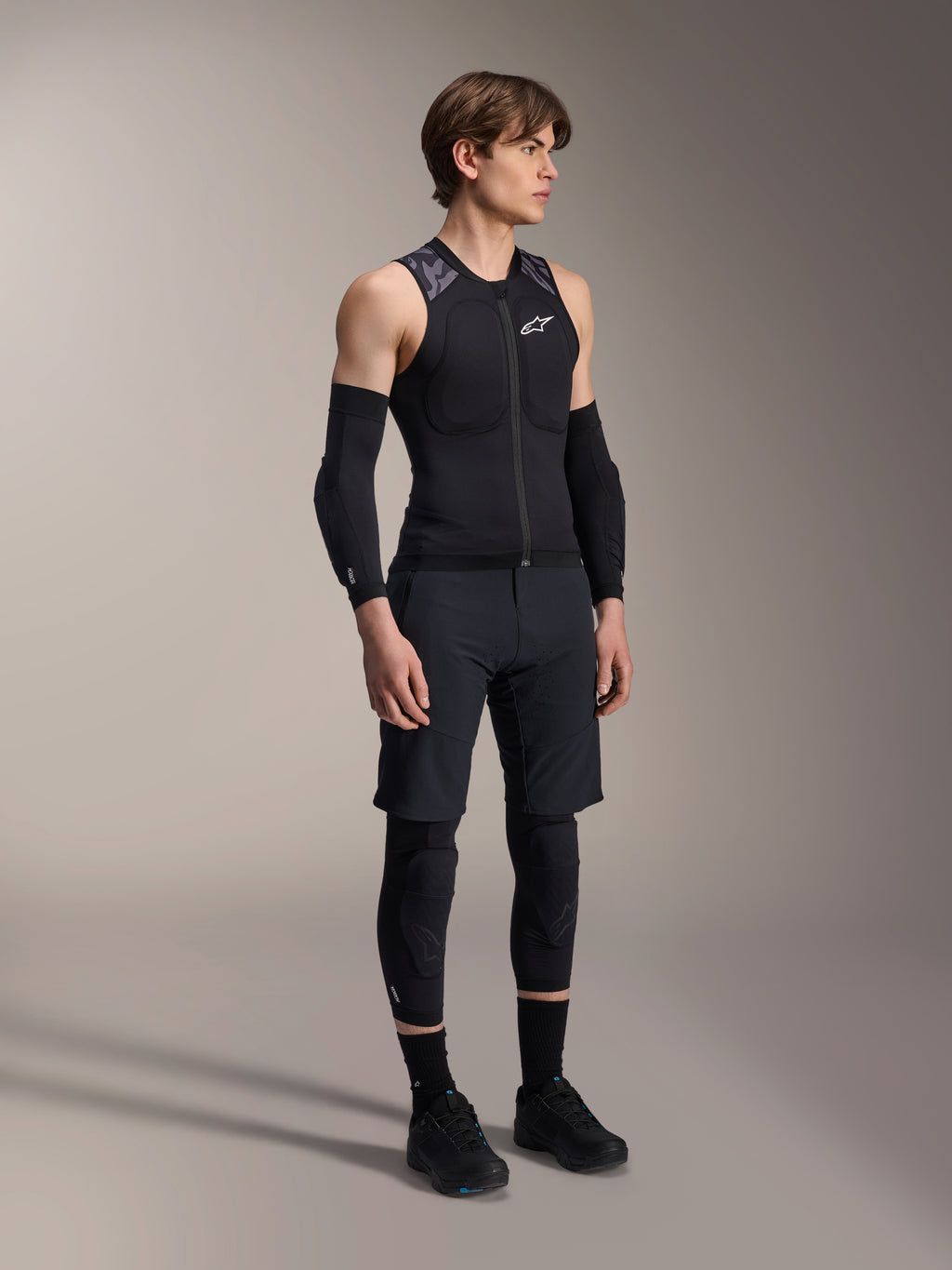 Alpinestars A-Motion Plasma skyddsväst, ärmlös svart och kamouflagemönstrad MTB-skyddsväst, helkroppsbild av modell som bär västen med ärmskydd och knäskydd, dragkedja framtill med vit Alpinestars-logotyp, högpresterande utrustning för mountainbikecykling