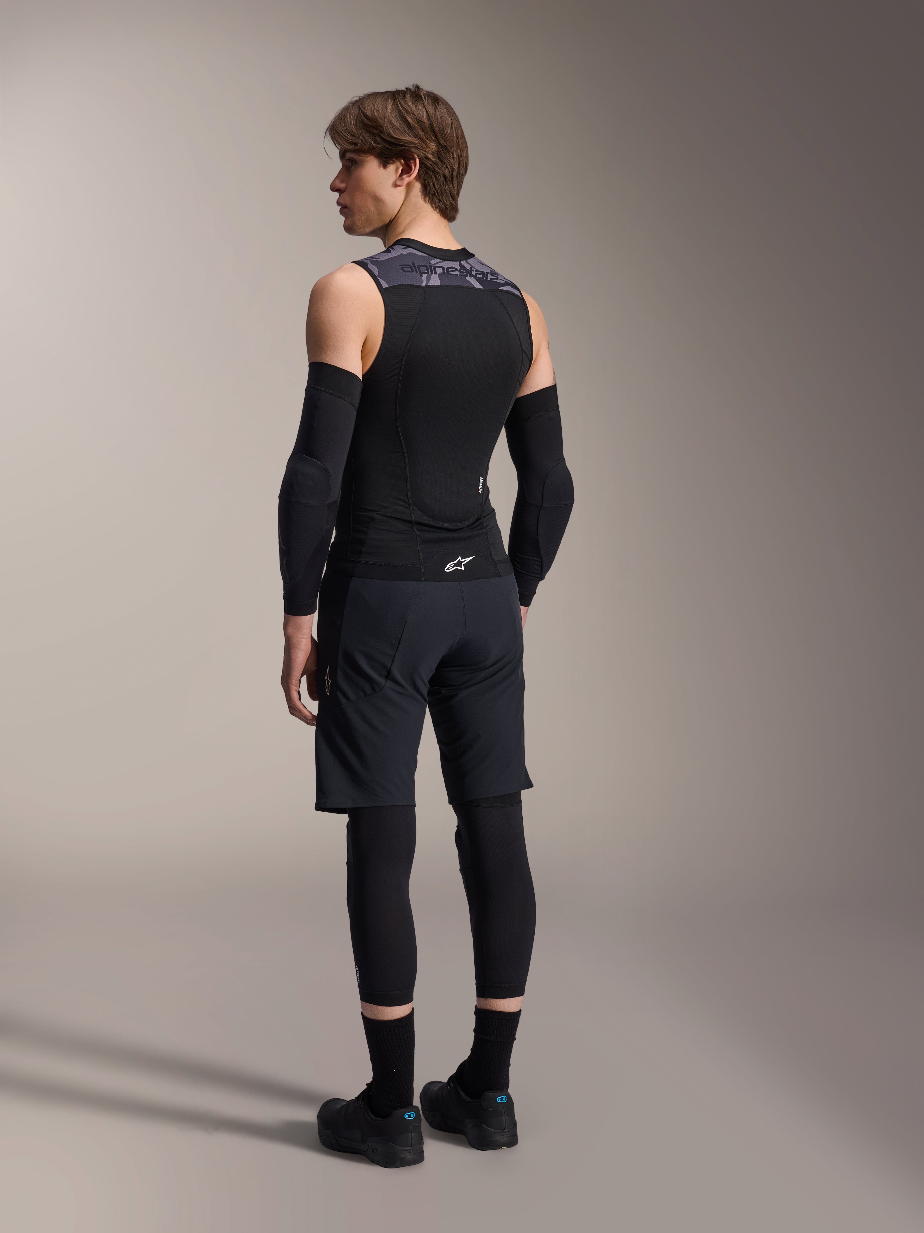 Manlig modell sedd bakifrån bär Alpinestars A-Motion Plasma skyddsväst, Black Camo, ärmlös mesh-design med integrerat ryggskydd, grått kamouflagemönster på axlarna, matchad med svarta armbågsskydd och mountainbike-shorts, professionell cykelutrustning