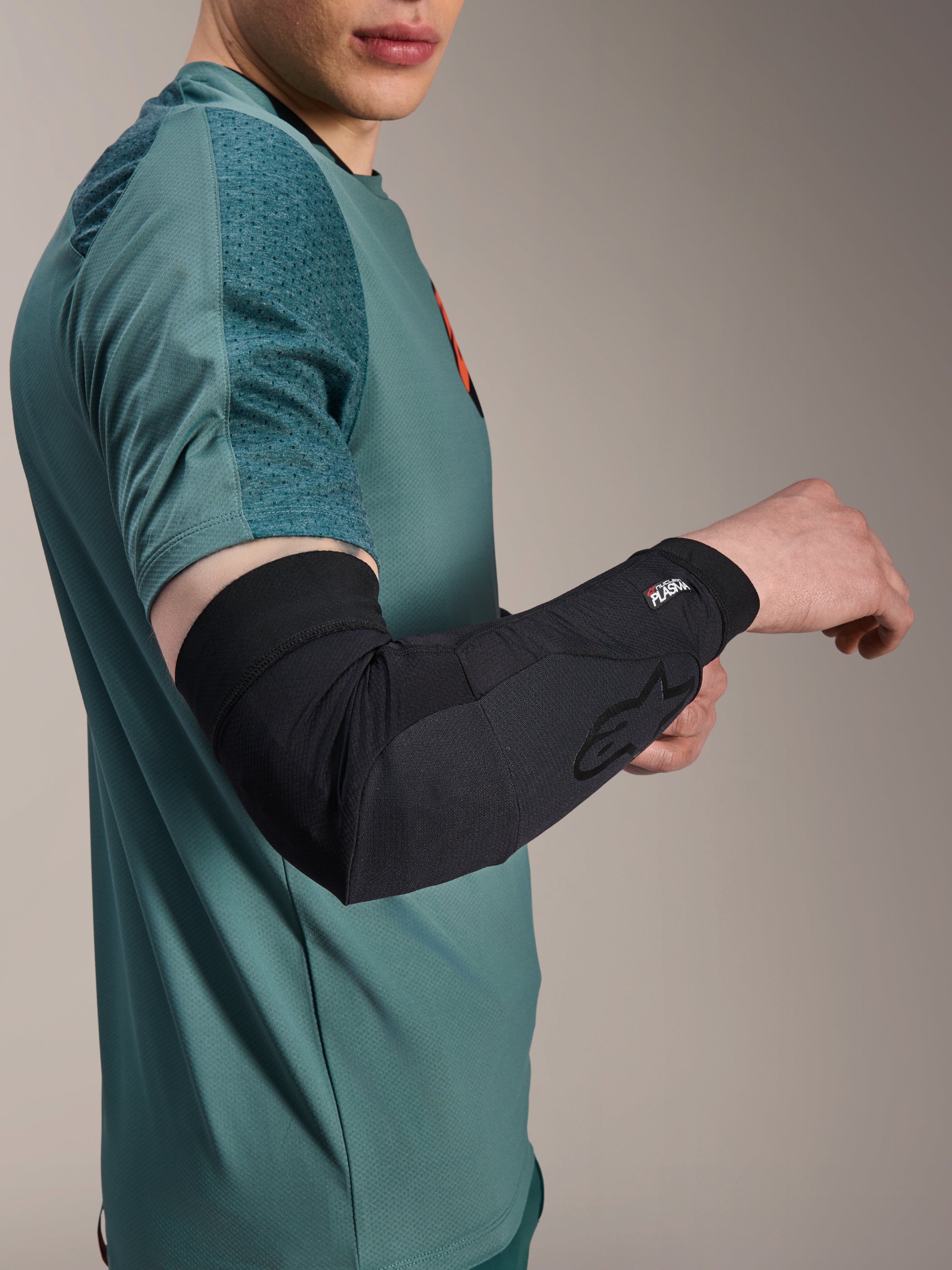 Alpinestars A-Motion Plasma armbågsskydd, svarta, bärs av en åkare i blågrön tröja, med Alpinestars-logotyp och Nucleon Plasma-märkning, smidig och ventilerande design för MTB-skydd