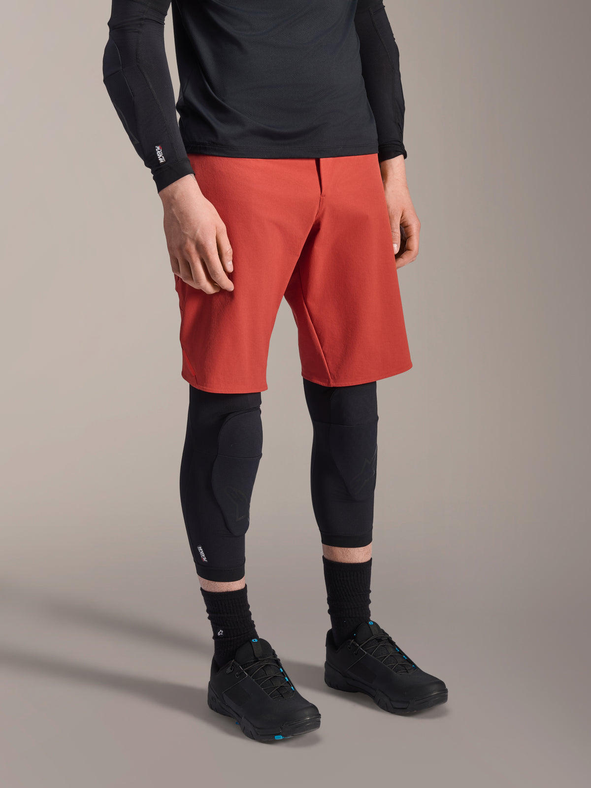 Alpinestars A-Motion Plasma knäskydd, MTB-knäskydd, svarta, bärs med röda shorts och svarta mountainbikeskor, lågprofils slip-on-design med konturerade skyddspaneler och ventilerande mesh.
