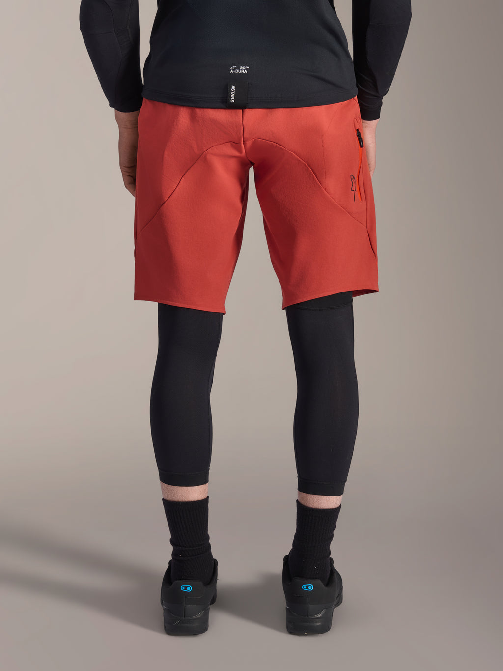 Alpinestars A-Motion Plasma knäskydd, svarta MTB-knäskydd, vy bakifrån burna under röda mountainbike-shorts med en svart cykeltröja och tekniska cykelskor, skyddsutrustning för mountainbikecykling