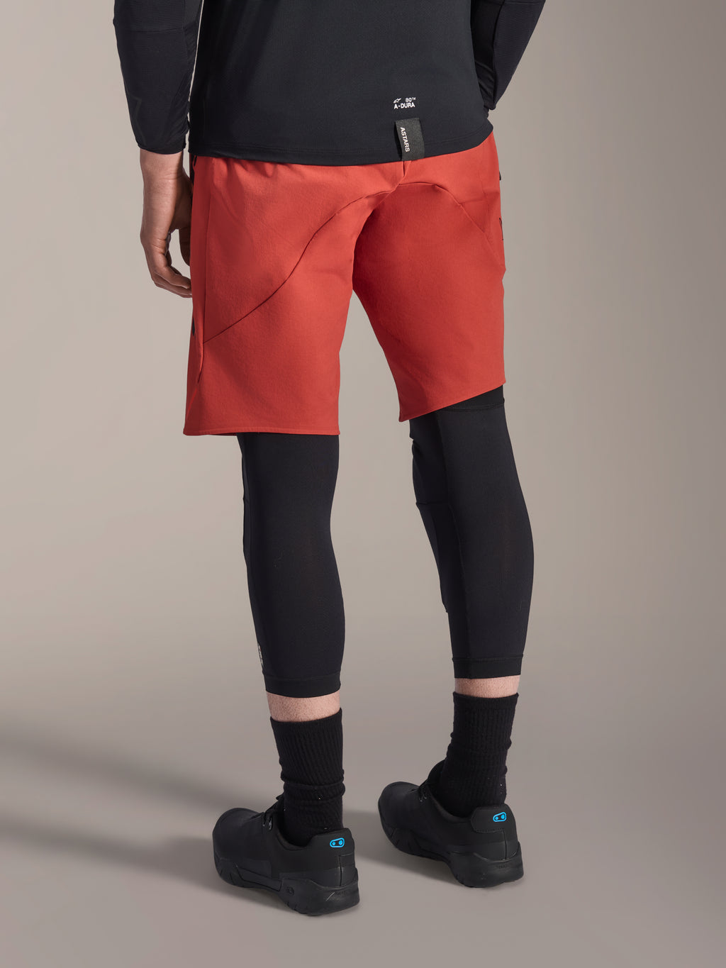 Alpinestars A-Motion Plasma knäskydd, svarta MTB-knäskydd, bärs med röda mountainbikeshorts och svarta cykelskor, vy bakifrån som visar den smidiga slip-on-designen för terrängcykling
