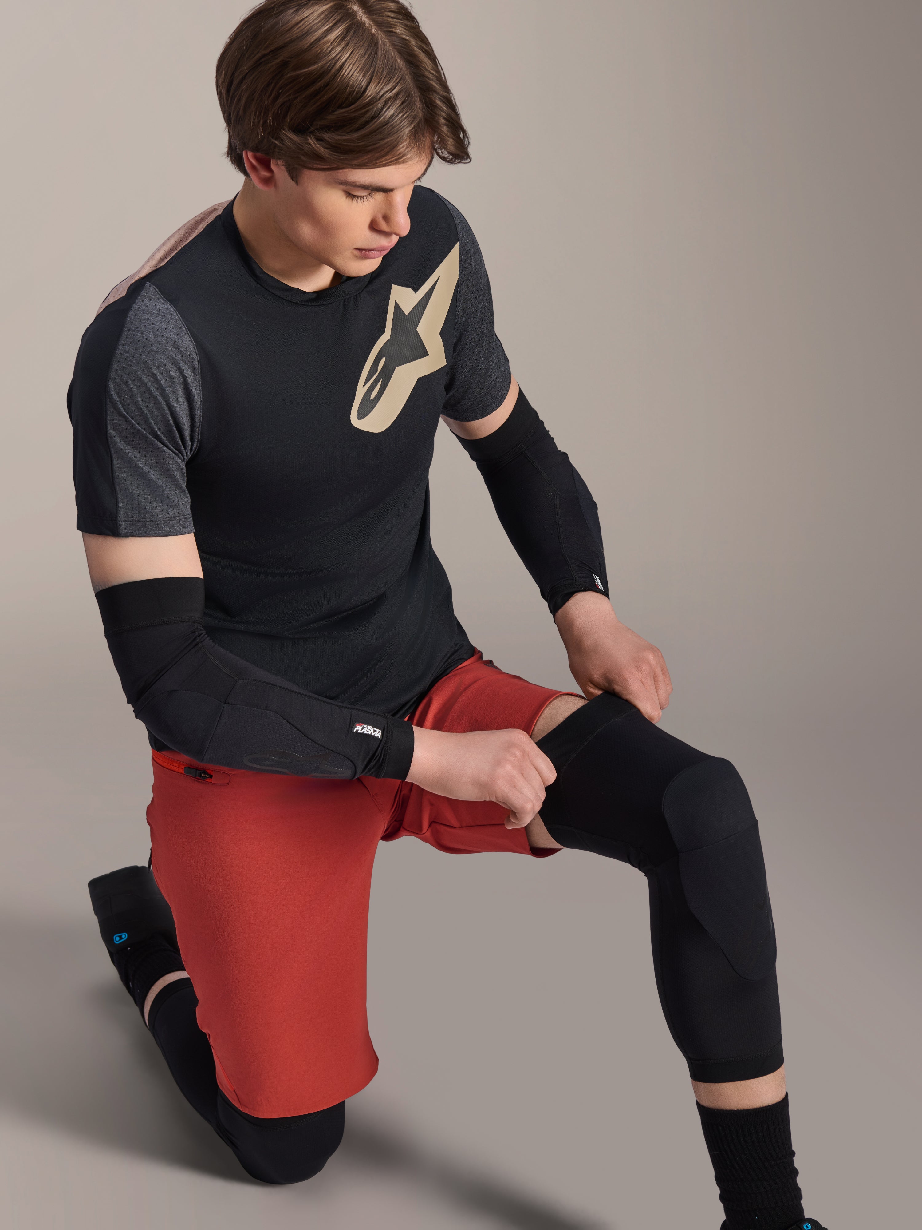 Alpinestars A-Motion Plasma Knee Sleeve, MTB-knäskydd, svart, bärs av en cyklist i knästående position, kombinerat med en svart och grå t-shirt med guldlogotyp, röda shorts och svarta armbågsskydd, professionell skyddsutrustning för mountainbike