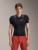 Manlig modell i Alpinestars A-Motion Plasma Protection Jacket - kortärmad, svart med grå kamouflagemönstrad axelvaddering, vit Alpinestars-logotyp på bröstet, kombinerad med röda byxor, kroppsnära MTB-skyddsjacka med dragkedja fram