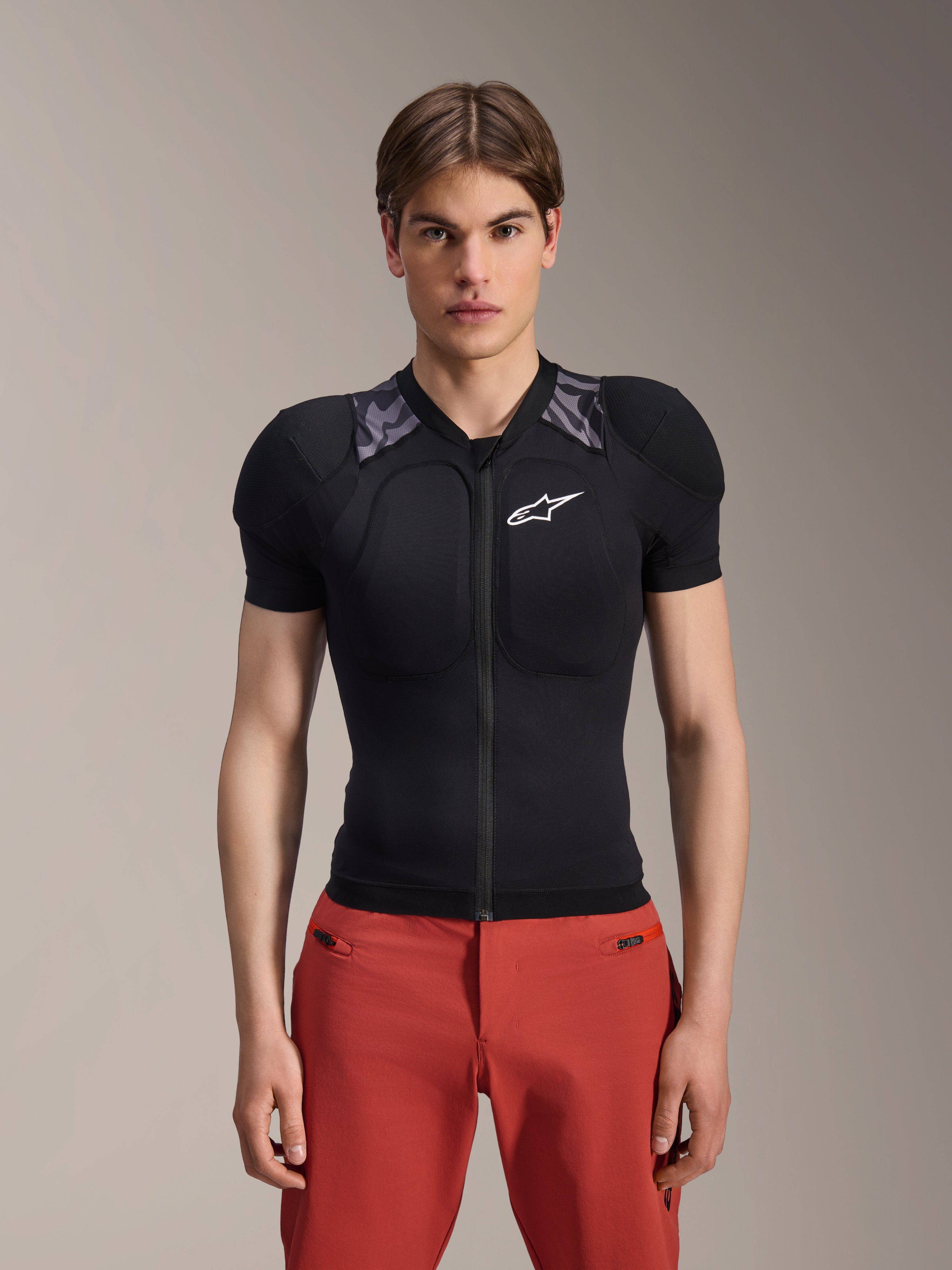 Manlig modell i Alpinestars A-Motion Plasma Protection Jacket - kortärmad, svart med grå kamouflagemönstrad axelvaddering, vit Alpinestars-logotyp på bröstet, kombinerad med röda byxor, kroppsnära MTB-skyddsjacka med dragkedja fram