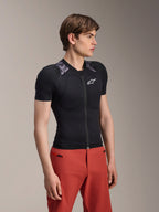 Manlig modell iklädd Alpinestars A-Motion Plasma skyddsjacka, kortärmad, Black Camo med grå kamouflagemönstrade axelpaneler, med integrerade axel- och bröstskydd, vit stjärnlogotyp på bröstet, ventilerande meshtyg, kombinerad med röda byxor