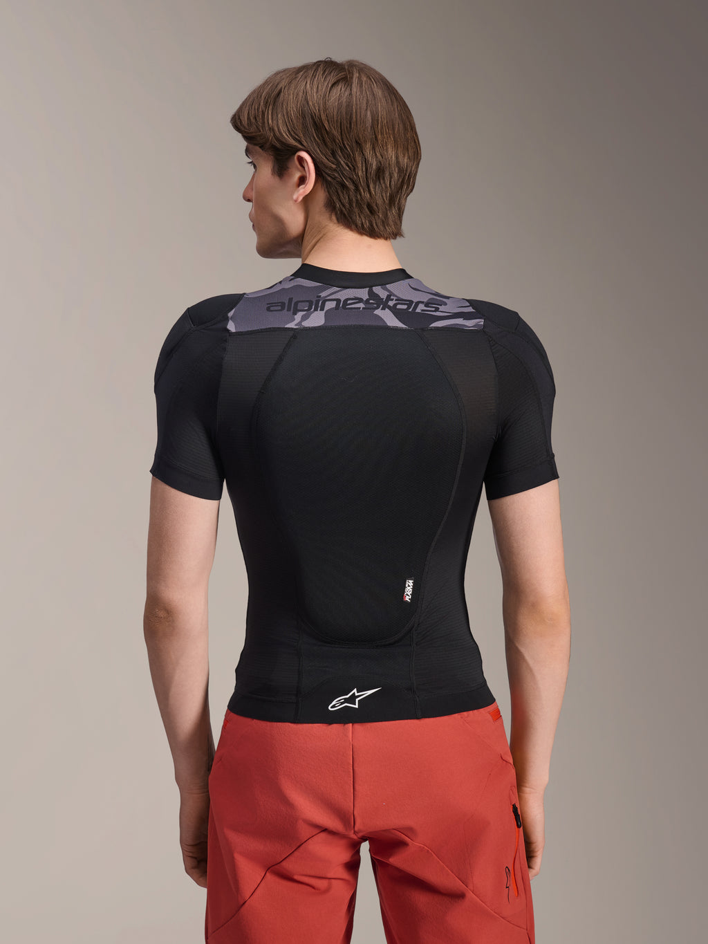 Alpinestars A-Motion Plasma skyddsjacka - kortärmad, svart med grå kamouflagedetaljer på övre ryggen, ryggvy som visar integrerat ryggskydd och vit Astars-logotyp i nederkanten, bärs med röda shorts, teknisk skyddsutrustning för mountainbike