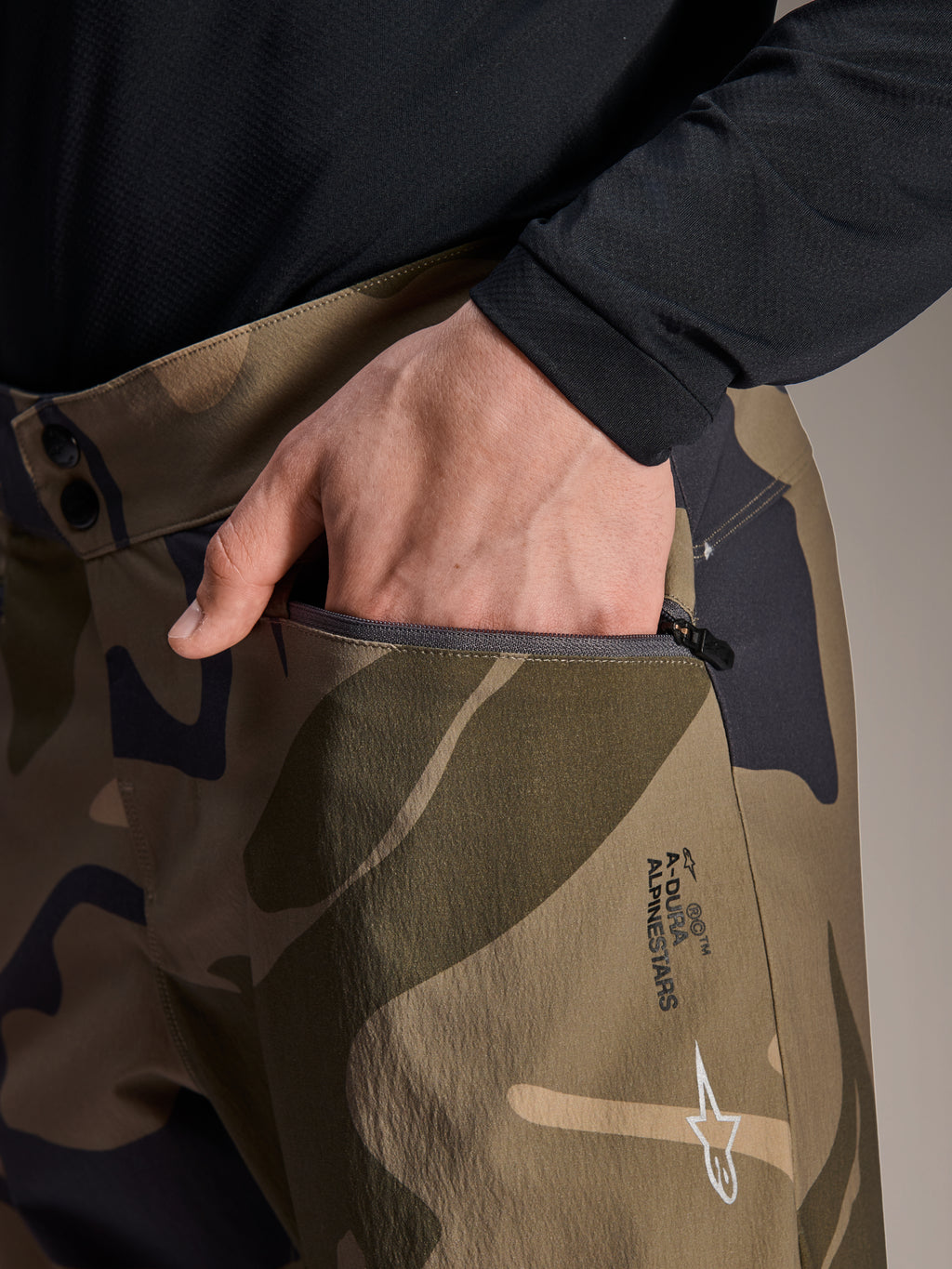 Närbild av Alpinestars A-Dura Camo Pant, bruna kamouflagemönstrade MTB-byxor, med en hand i en blixtlåsförsedd ficka, dubbel knäppning i midjan och Alpinestars-logotyp på låret.