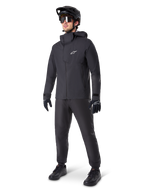 Manlig mountainbike-cyklist klädd i svarta Alpinestars A-Dura Waterproof Pants, matchande svart vattentät jacka med huva, svart MTB-hjälm med visir, mörka cykelglasögon och svarta handskar, komplett professionellt utrustningsset för terrängcykling i alla väder