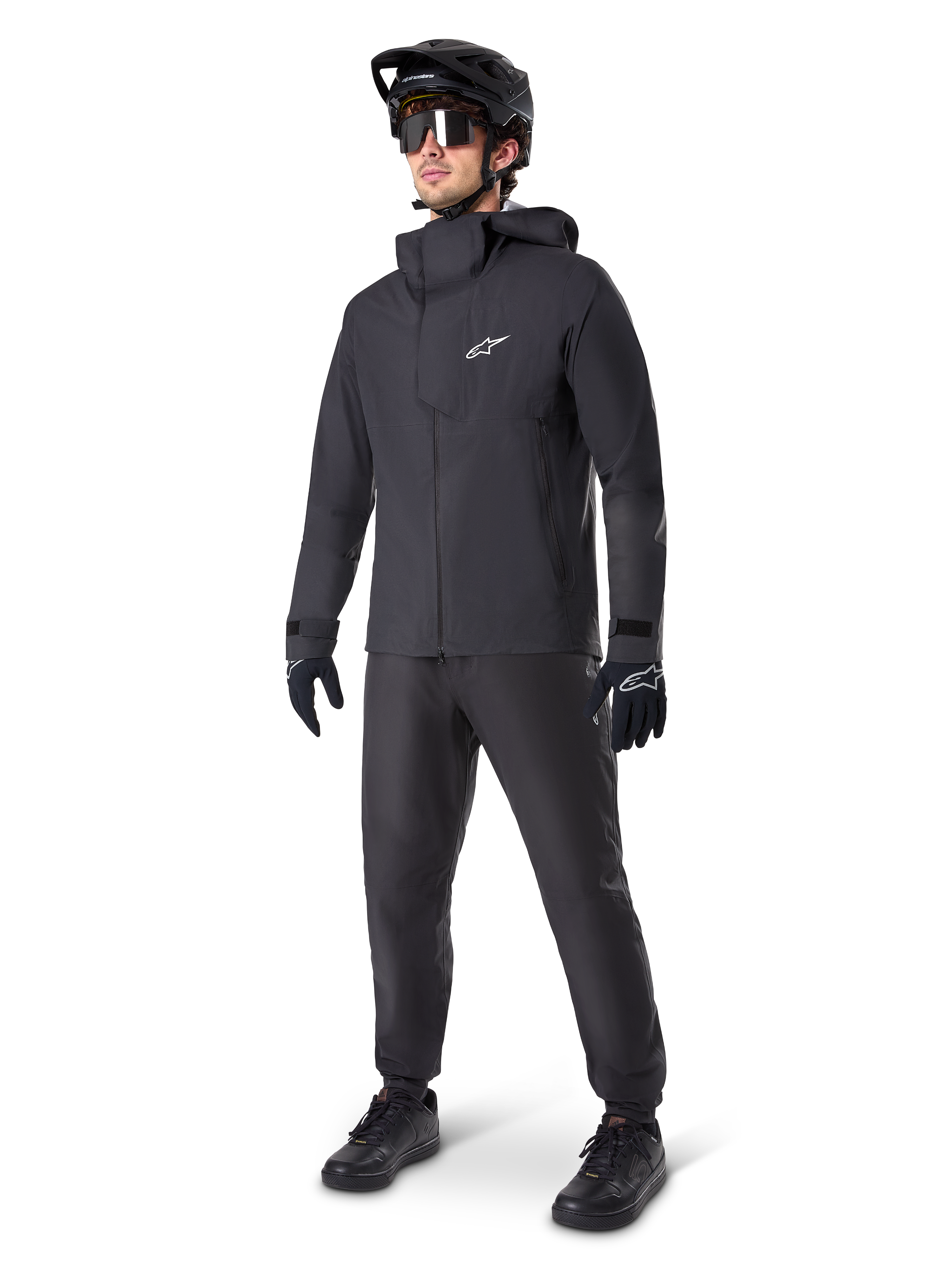 Manlig mountainbike-cyklist klädd i svarta Alpinestars A-Dura Waterproof Pants, matchande svart vattentät jacka med huva, svart MTB-hjälm med visir, mörka cykelglasögon och svarta handskar, komplett professionellt utrustningsset för terrängcykling i alla väder