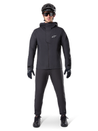 Alpinestars A-Dura Waterproof Pants, svarta MTB-byxor, bärs som en del av en komplett helsvart mountainbike-utrustning inklusive en teknisk vattentät jacka, svart hjälm, solglasögon och handskar, designade för terrängkörning i vått väder.
