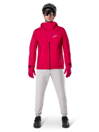 Alpinestars A-Dura Waterproof Pants, MTB-byxor, Titanium Gray, bärs med en starkröd jacka med huva, röda handskar och en svart mountainbikehjälm, professionell vattentät cykelutrustning för våta terrängförhållanden