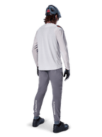 Alpinestars A-Aria Elite-byxor, MTB-byxor, mörkgrå, vy bakifrån av en cyklist i en hel outfit, vit Alpinestars-logotyp på underbenen, prestandapassform med laserperforeringar, matchad med en ljusgrå långärmad tröja och grå MTB-hjälm