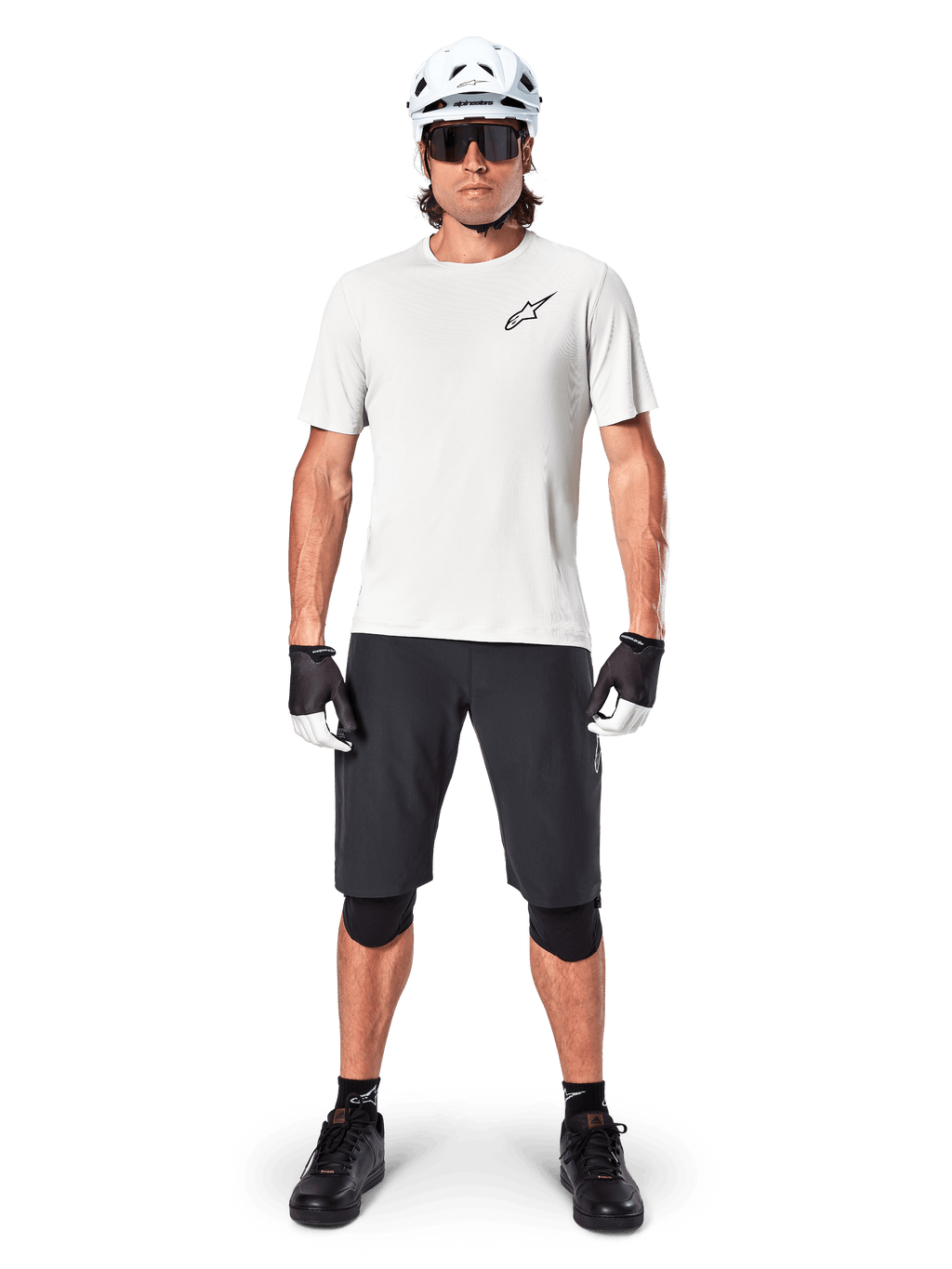Manlig mountainbikecyklist i Alpinestars A-Aria Shorts, svarta, tillsammans med en vit funktionströja, vit hjälm och svarta knäskydd, professionell utrustning för terrängcykling, helbild framifrån