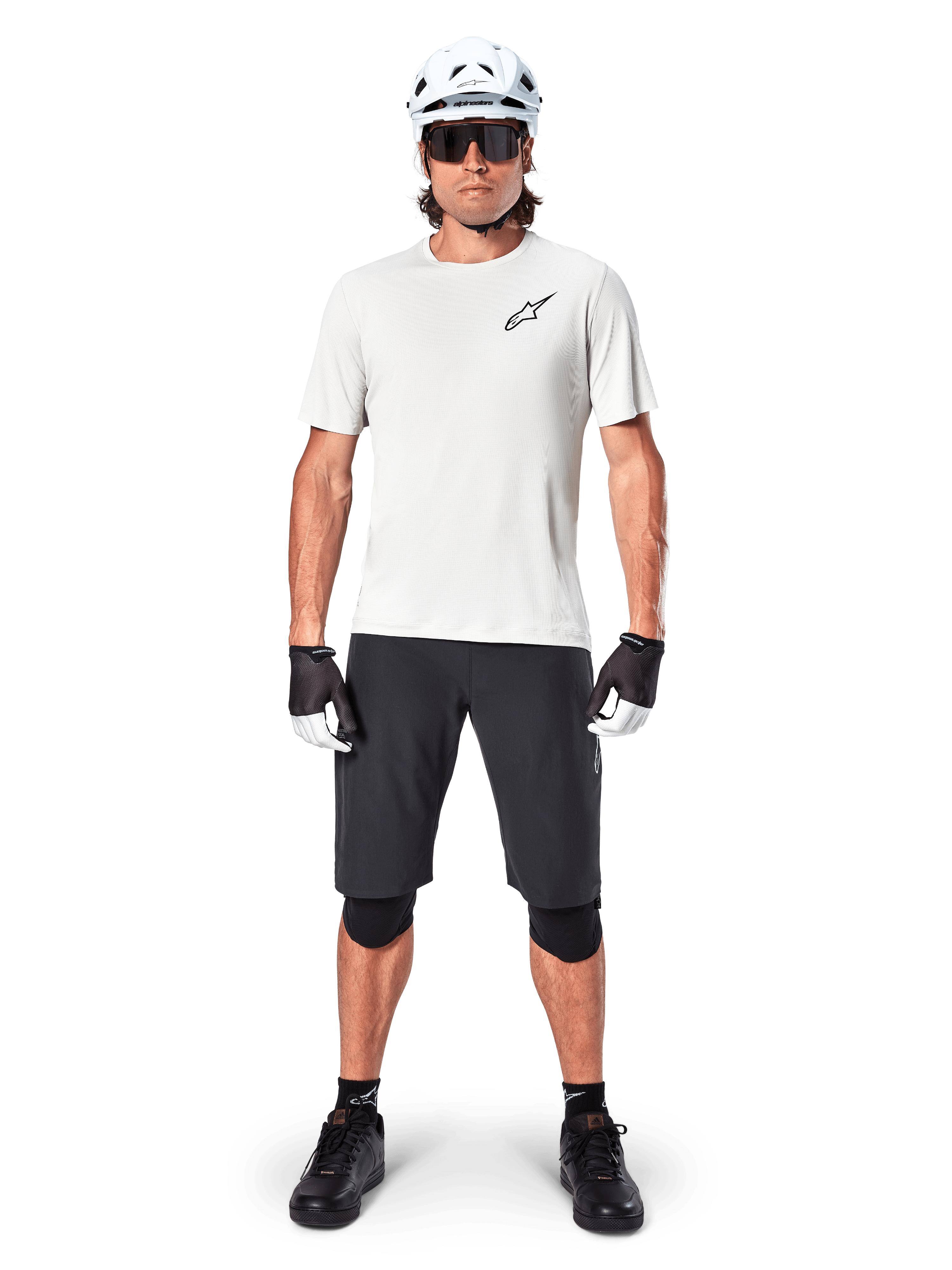 Manlig mountainbikecyklist i Alpinestars A-Aria Shorts, svarta, tillsammans med en vit funktionströja, vit hjälm och svarta knäskydd, professionell utrustning för terrängcykling, helbild framifrån