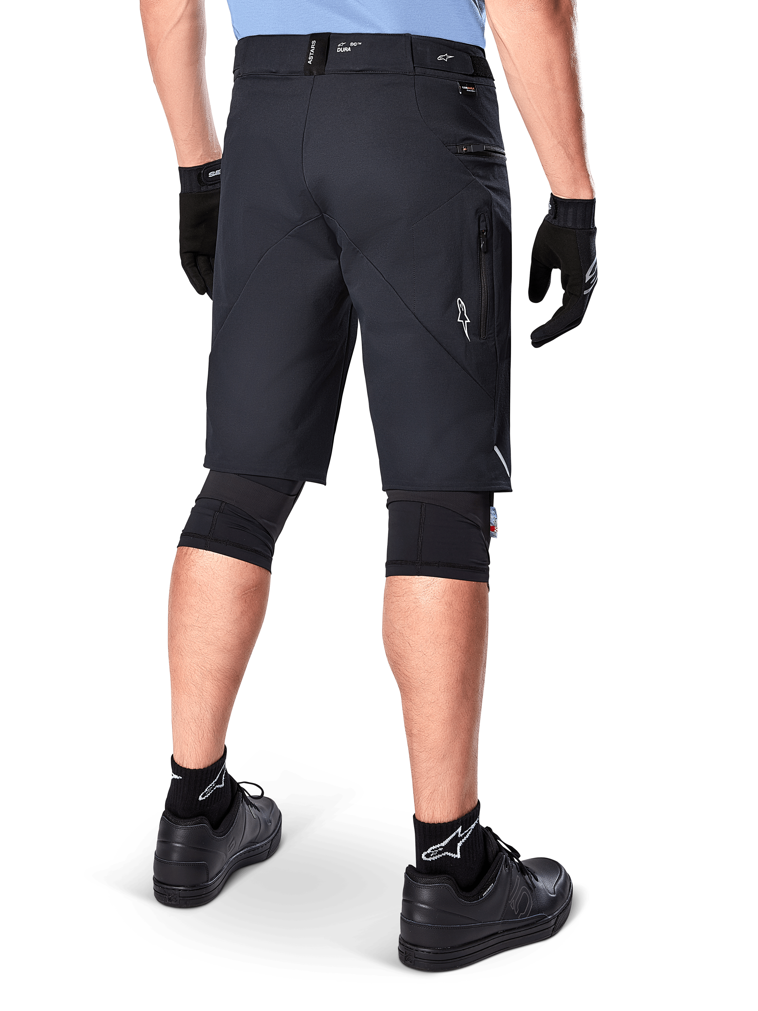 Alpinestars A-Dura Elite Shorts, Black, rear view of a rider wearing mountain bike gear including black knee pads, gloves, and socks with white logos, featuring zippered side pockets and reflective accents on a durable Cordura fabric.