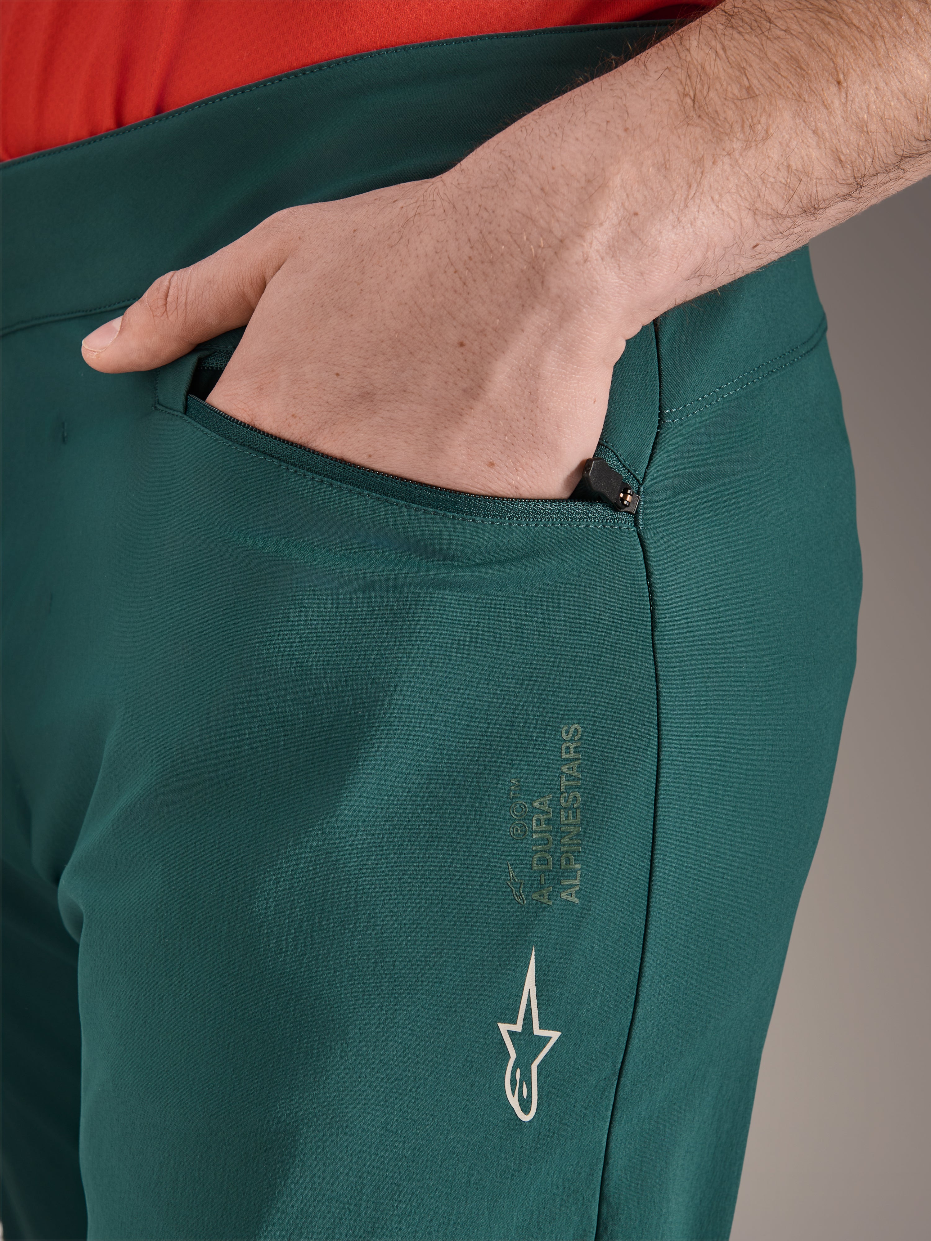 Alpinestars A-Dura byxor i färgen Dark Pine, närbild på blixtlåsförsedd ficka med hand, med vit Alpinestars-logotyp och text på låret, slitstarka MTB-cykelbyxor